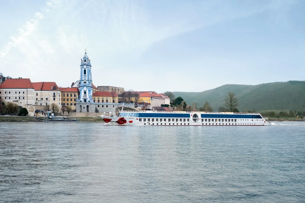 A white river cruise ship sails along a calm river, with a picturesque town featuring a striking blue and white church tower and red-roofed buildings in the background, set against rolling green hills.