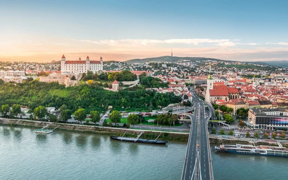 A scenic view of Bratislava, showcasing the historic castle on a hill surrounded by lush greenery, with a river and bridge in the foreground. The cityscape includes a prominent church with a tall spire and a backdrop of rolling hills under a clear sky.