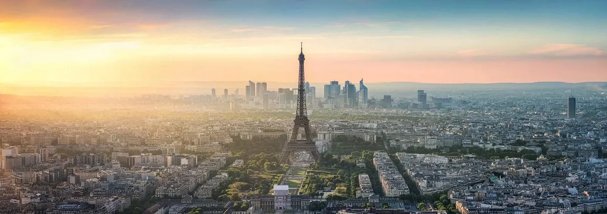 A panoramic view of Paris at sunset, featuring the Eiffel Tower prominently in the center, surrounded by a sprawling cityscape with a mix of historic and modern buildings, as the sky transitions from a warm golden hue to a soft blue.