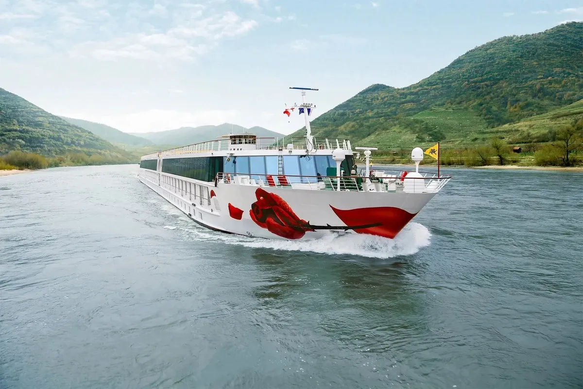 A river cruise ship with a striking red rose design on its bow sails through a scenic landscape, surrounded by lush green hills under a clear blue sky.
