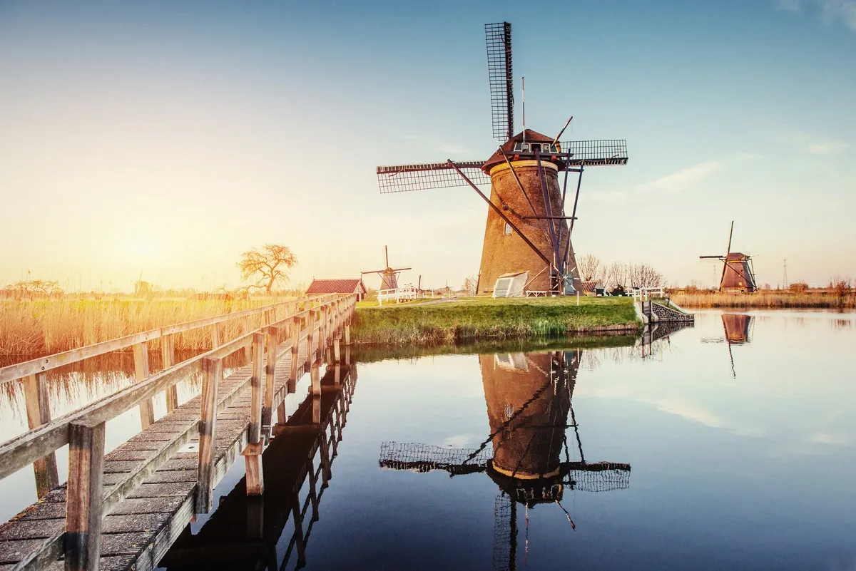 A picturesque scene of a traditional windmill reflected in calm water, with a wooden walkway leading towards it. The setting sun casts a warm glow over the landscape, highlighting the clear blue sky and distant windmills in the background.