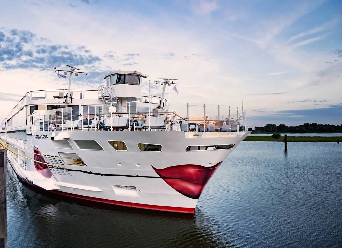 A white cruise ship with a distinctive red lips design on the bow is docked in calm waters under a partly cloudy sky. The vessel, featuring multiple decks and a modern bridge, is moored near a grassy shoreline.