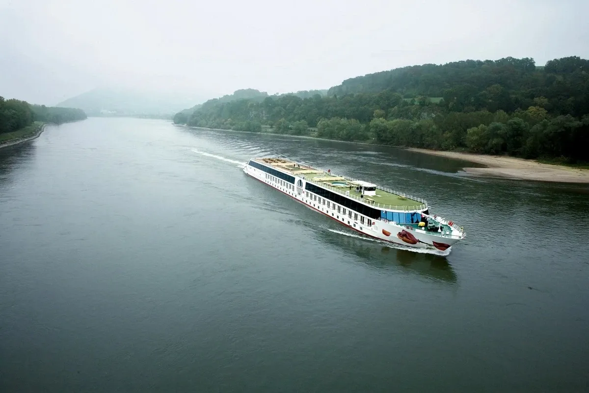 A white river cruise ship sails along a wide, calm river, bordered by lush green trees and a sandy shore under a misty sky.
