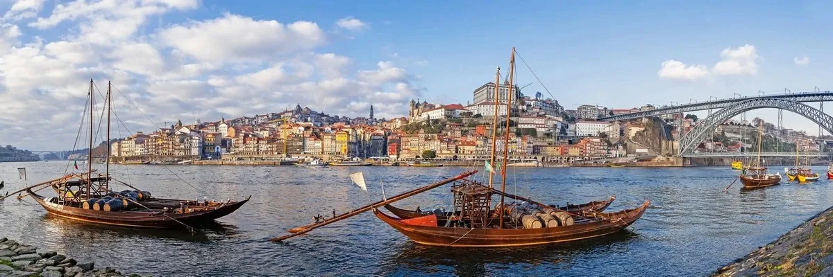 Traditional wooden boats with barrels float on a river, set against a picturesque cityscape with colorful buildings and a prominent arched bridge under a partly cloudy sky.