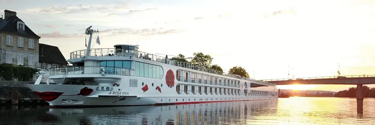 A river cruise ship named A-Rosa Viva is docked near a charming riverside building, with a bridge and a picturesque sunset in the background. The ship features a distinctive design with red lips and rose motifs on its hull.