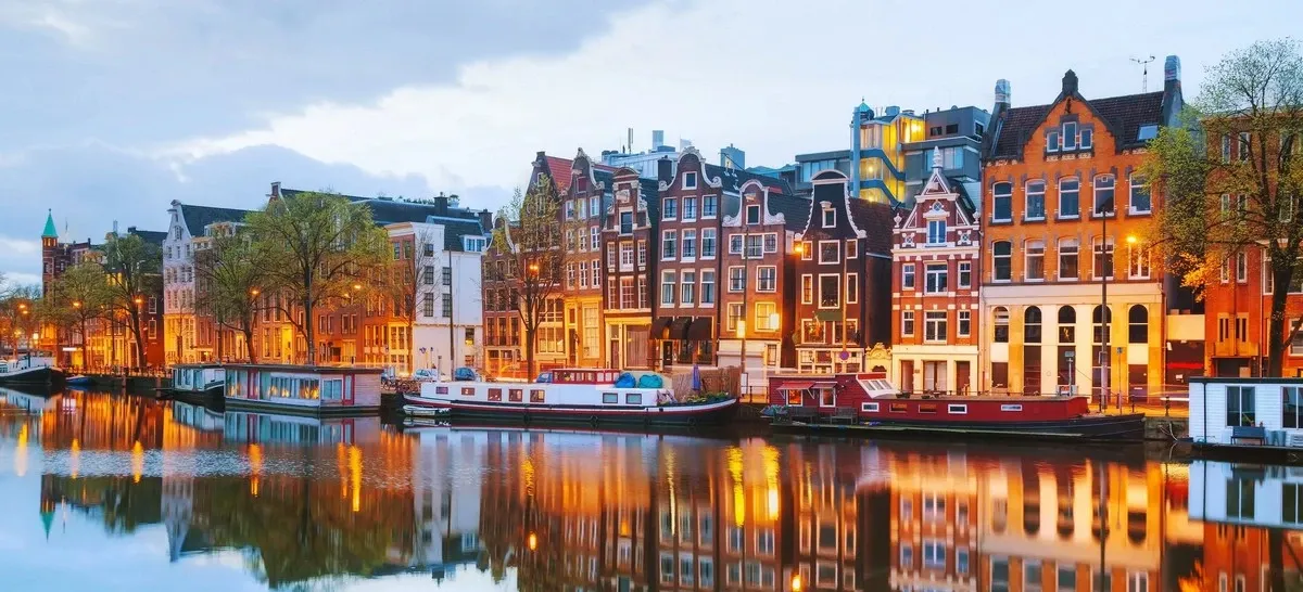 A row of charming, narrow buildings lines a canal in Amsterdam, their warm lights reflecting beautifully in the water. The scene is peaceful, with houseboats moored along the canal and trees adding a touch of greenery.