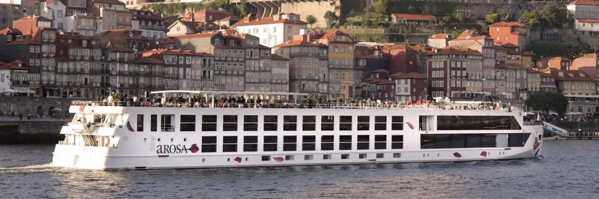 A white A-ROSA river cruise ship sails along a river with a picturesque backdrop of densely packed, colorful buildings on a hillside, bathed in warm sunlight.