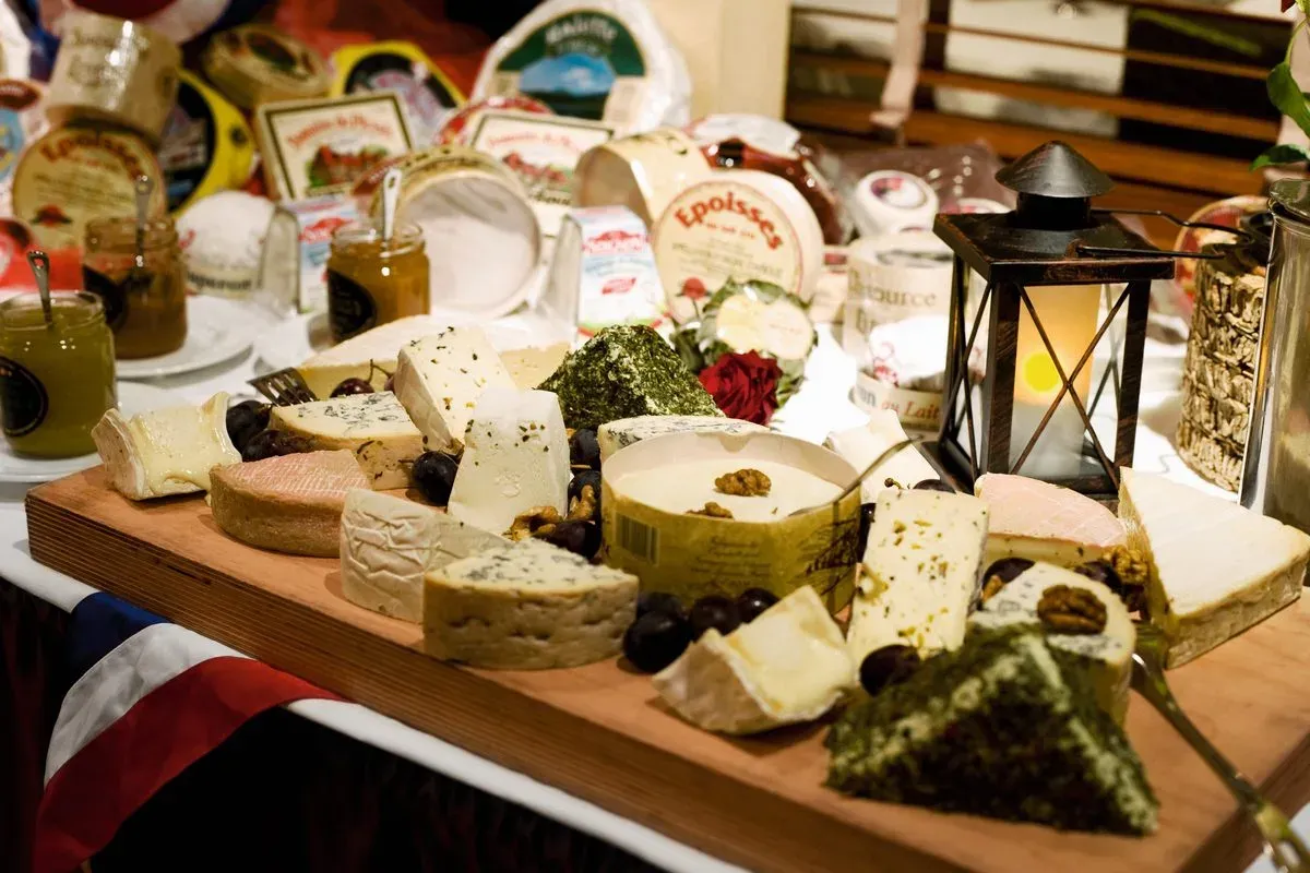 A wooden board displays an assortment of cheeses, including varieties with herbs and nuts, surrounded by dark grapes. In the background, jars of jam and various cheese packages are visible, along with a decorative lantern casting a warm glow.