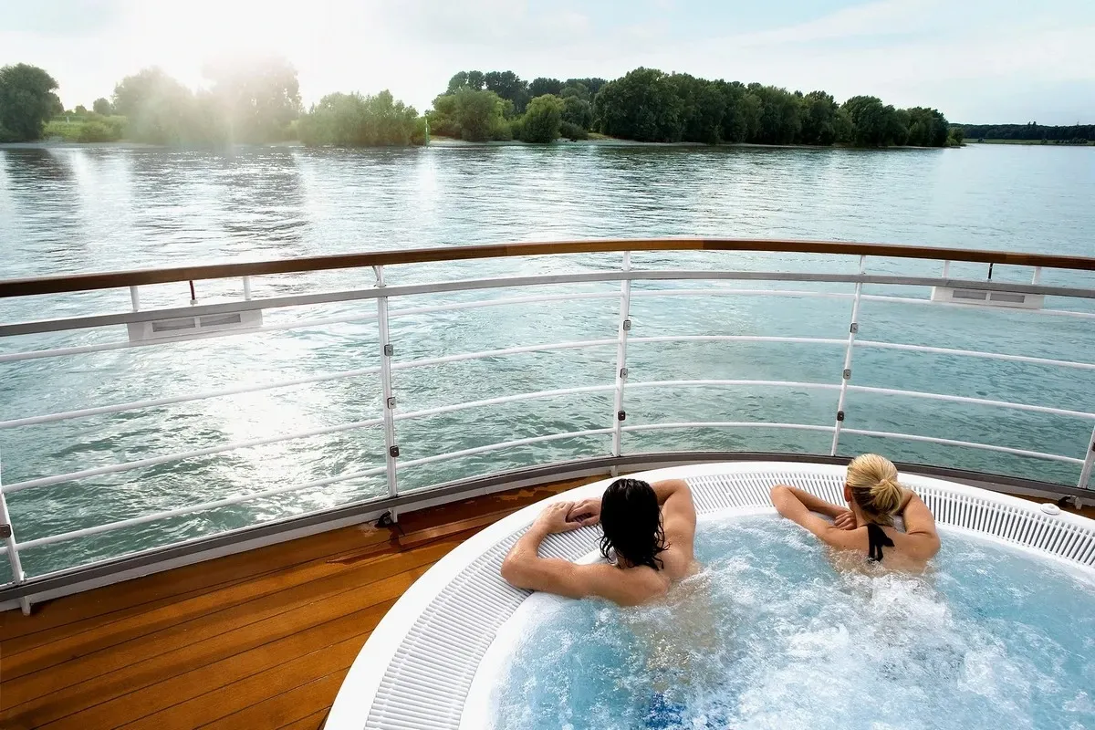 A couple relaxes in a bubbling hot tub on a cruise ship's deck, overlooking a serene river with lush green trees lining the distant shore under a bright sky.