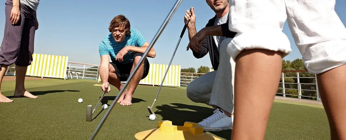 Three people are playing mini-golf on a sunny day, with one person in a blue striped shirt crouching to line up a putt. Another person in white pants and sneakers is standing nearby, focusing on the game.
