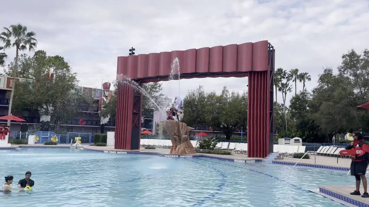 A swimming pool featuring a large red arch with a water fountain spraying into the pool. The setting includes trees and lounge chairs, with a cloudy sky overhead.