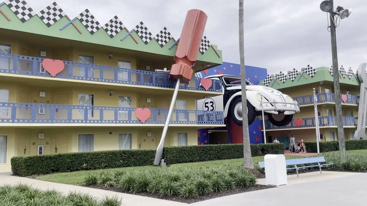 A colorful hotel facade features a giant classic car with the number 53, seemingly driving out of the building. Large decorative elements, like a massive screwdriver and red hearts, adorn the bright yellow and blue exterior. There's a checkered pattern along the roofline, and a few people walk along the pathway in front.