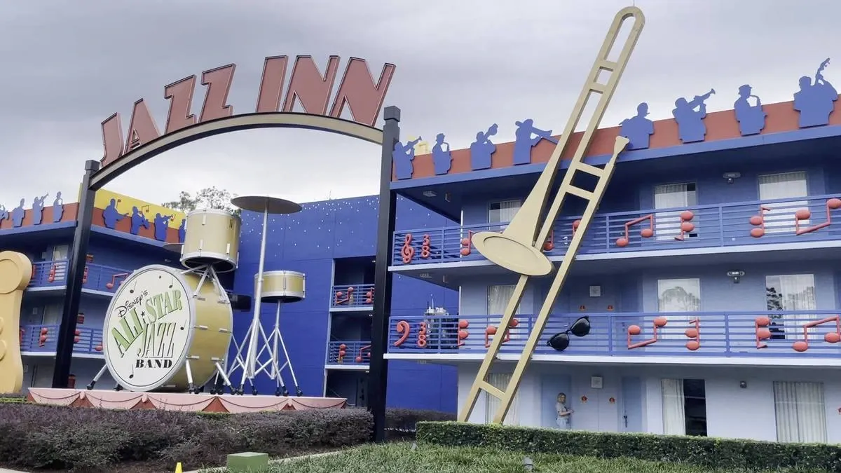 The image shows the exterior of the Jazz Inn at Disney's All-Star Music Resort, featuring a large drum set and trombone sculpture. The building is painted blue with musical notes and silhouettes of musicians decorating the facade. The sign above reads "JAZZ INN" in bold letters.