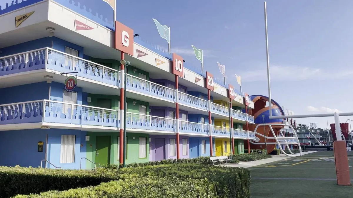 A colorful hotel building features three stories with blue, green, yellow, and purple doors, each level marked with large numbers and letters. The exterior is adorned with football-themed decorations, including a giant helmet and goalpost.