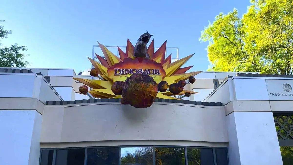 The entrance to the "DINOSAUR" attraction features a dramatic sign with a fiery explosion design and a large meteor at the center, set against a bright blue sky with green trees surrounding the building.