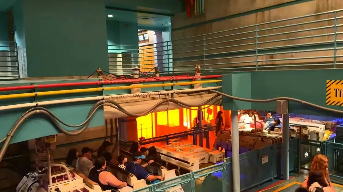 People are seated in a theme park ride featuring white vehicles resembling boats, set in an industrial-themed area with colorful pipes and orange lighting.
