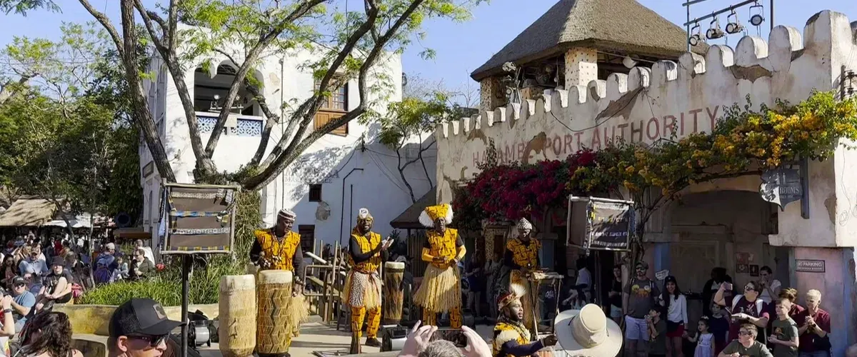 Performers in vibrant traditional attire play drums in front of a lively crowd at an outdoor venue with a rustic building and lush greenery.