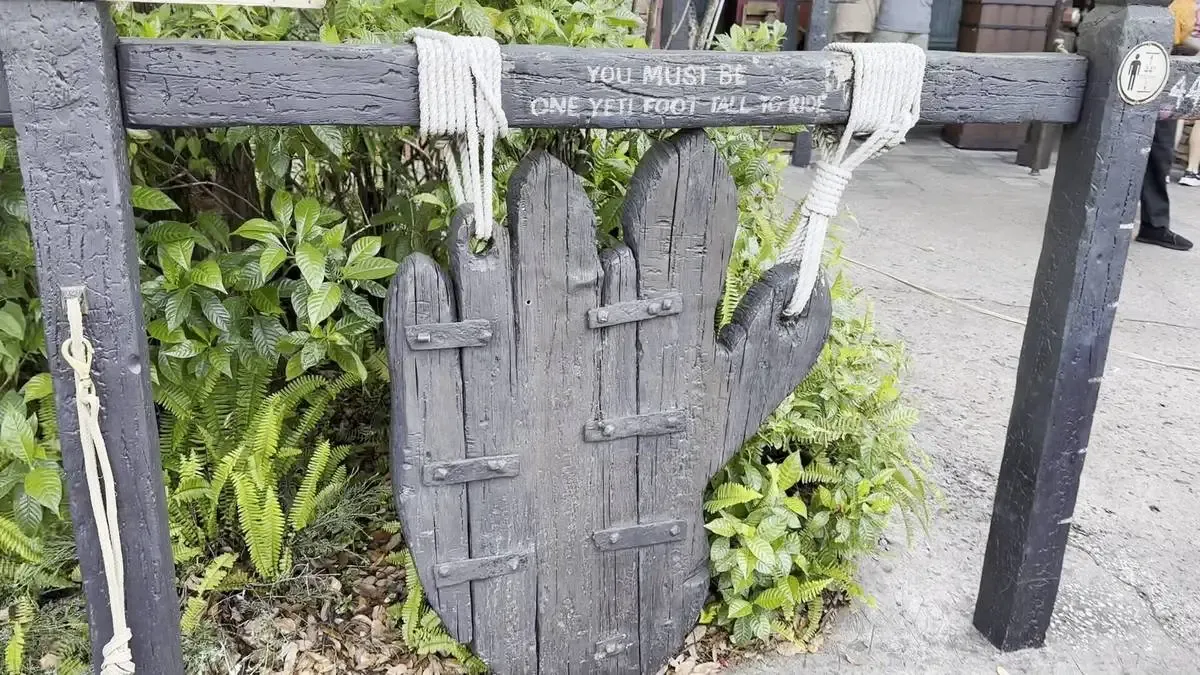 A wooden height requirement sign shaped like a large hand is displayed in front of lush green foliage. The sign humorously states, "You must be one Yeti foot tall to ride," with ropes securing it to the frame.