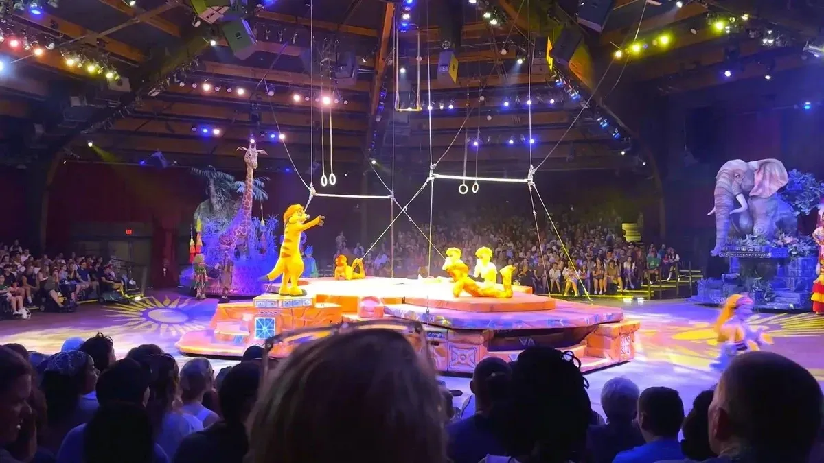 A vibrant stage performance featuring characters in animal costumes takes place under colorful lights, with a large audience watching. The set is decorated with jungle-themed elements, including a large elephant figure and a platform with ropes and rings hanging above.