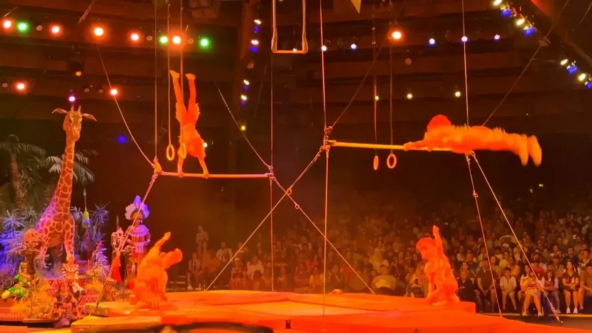 Acrobats perform a dynamic routine on trapeze bars under colorful stage lights, with one hanging upside down and another mid-air. A giraffe prop and vibrant costumes add to the lively atmosphere, while a captivated audience watches in the background.