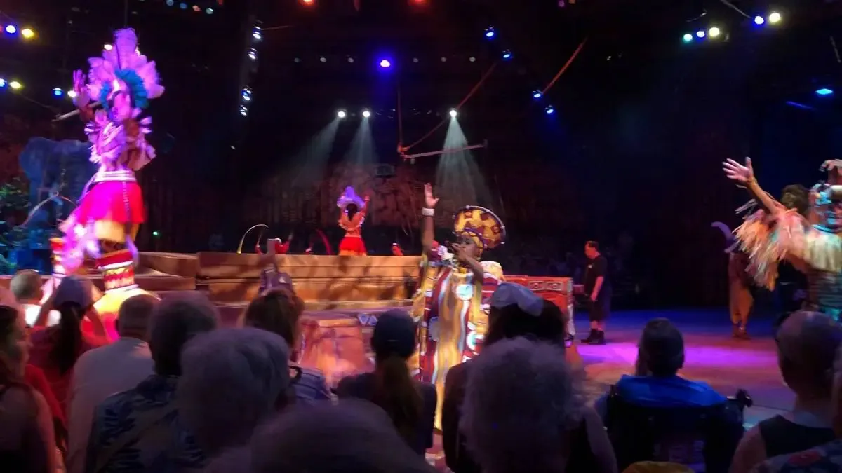 Performers in vibrant costumes with feathered headdresses and colorful outfits entertain a seated audience in a lively, illuminated stage setting.