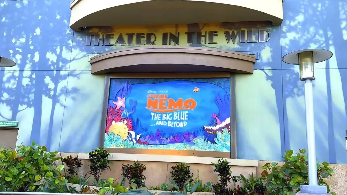 The entrance to "Theater in the Wild" features a colorful poster for "Finding Nemo: The Big Blue and Beyond," surrounded by lush greenery and a lamppost, set against a backdrop of painted trees.