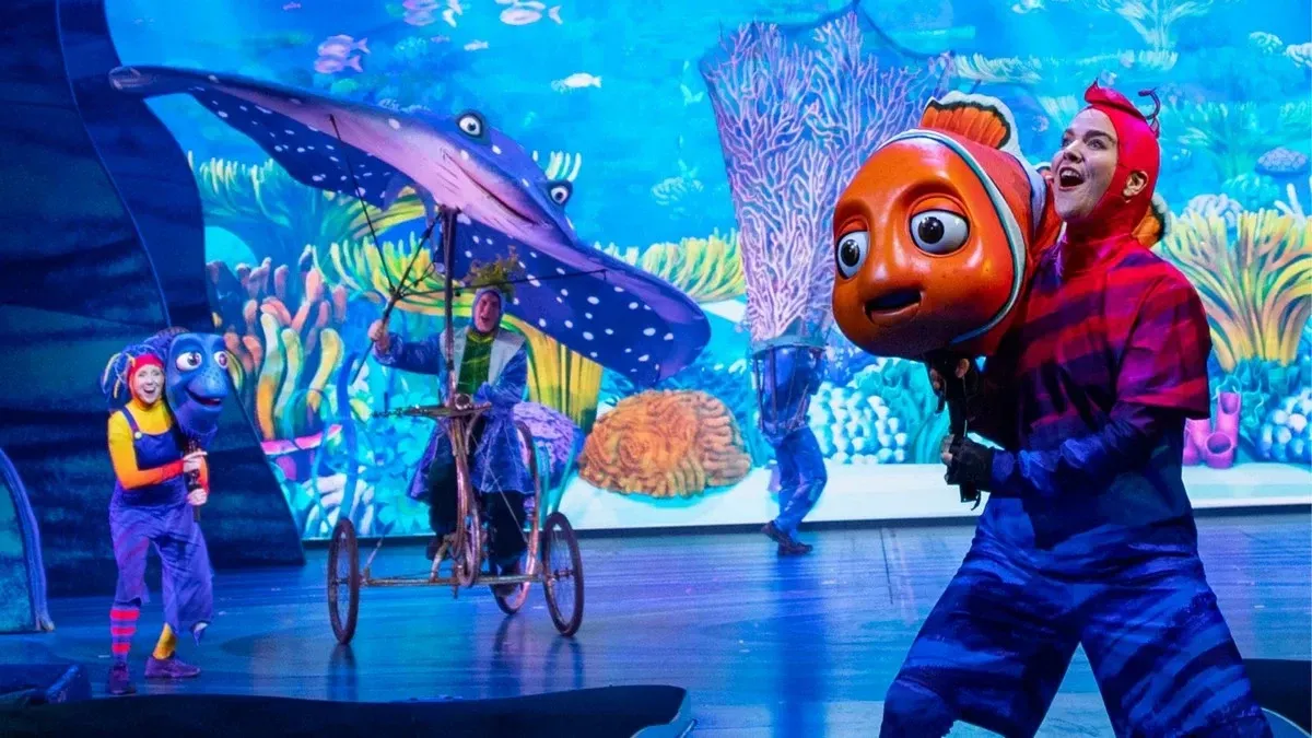 Performers on stage bring "Finding Nemo" to life with colorful costumes and large character puppets, including a vibrant clownfish and a spotted stingray, set against an underwater backdrop.