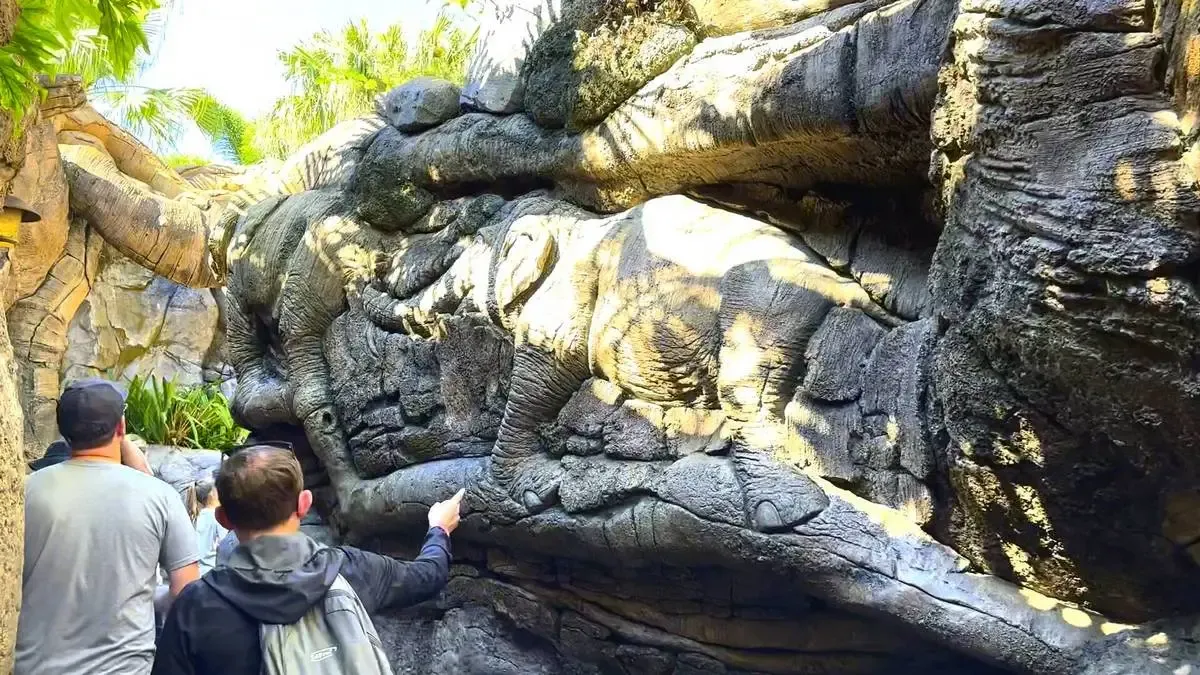 People are observing a rock formation that resembles an elephant, with detailed textures and shading, in a sunlit outdoor setting surrounded by greenery.