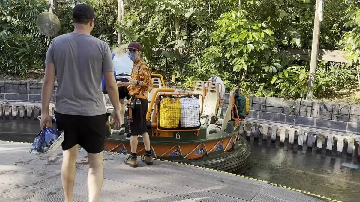 A person in a gray shirt and black shorts walks towards a river ride where another person assists passengers onto a circular raft. The raft, marked with the number 6, is surrounded by lush greenery and bordered by a stone wall.