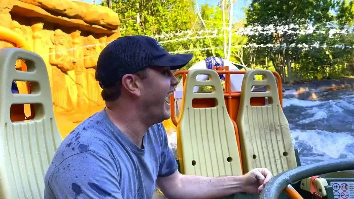 A man wearing a wet gray shirt and a dark cap is enjoying a water ride, gripping the railing of a raft with excitement. The ride features orange and beige seats, with splashing water and greenery in the background, creating a lively amusement park atmosphere.