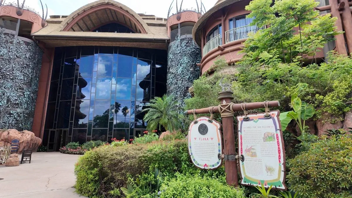 The image shows a large, architecturally unique building with a curved, thatched roof and a facade of tall glass windows reflecting a blue sky with clouds. The entrance is flanked by tropical greenery and informational signs.