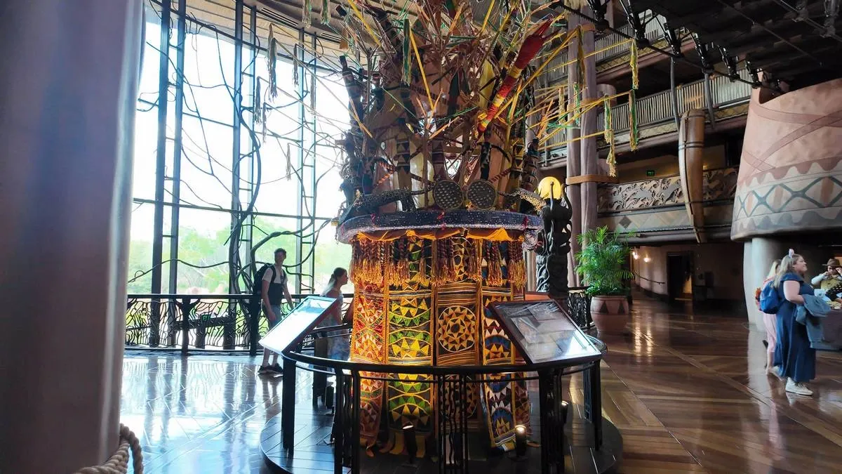 A colorful display featuring intricate patterns and woven materials stands prominently in a spacious, warmly lit room with large windows. People are casually walking around, taking in the decor and atmosphere.