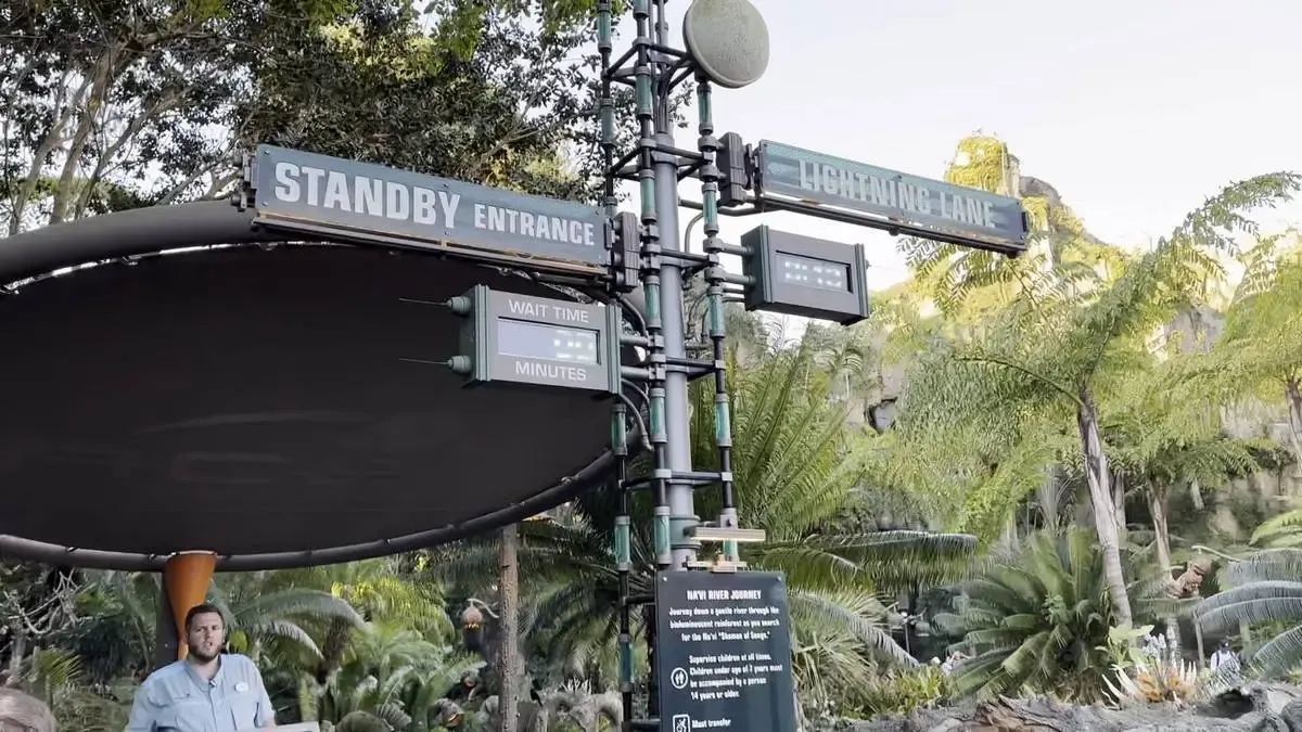 A theme park entrance sign shows "Standby Entrance" and "Lightning Lane" with digital displays for wait times. Lush greenery surrounds the area, and a person stands nearby, enhancing the adventurous atmosphere.