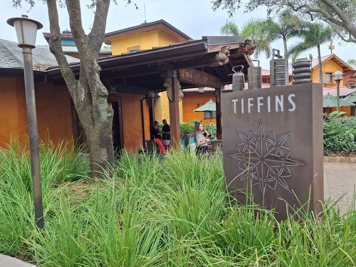 The image shows the entrance to "Tiffins", a restaurant with a rustic exterior surrounded by lush greenery. A prominent sign with the name of the restaurant and a compass design stands in the foreground. In the background, people can be seen sitting under umbrellas, enjoying the outdoor setting.