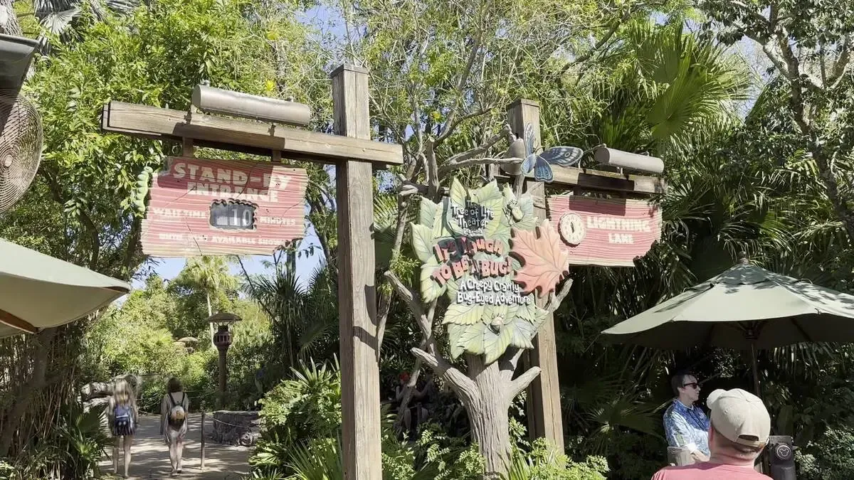 Entrance to the "It's Tough to Be a Bug!" attraction, featuring a rustic sign with colorful leaf decorations, surrounded by lush greenery. Visitors are walking towards the attraction under a sunny sky, with a person sitting under a green umbrella to the right.