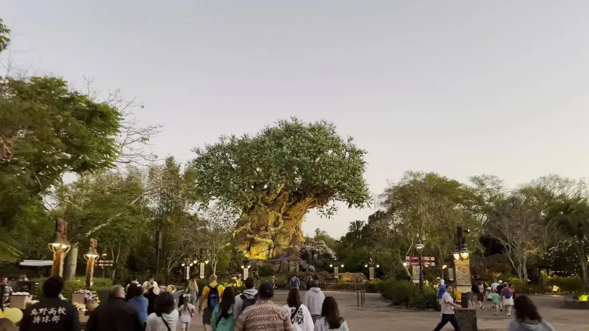 A group of people walk toward a large, intricately designed artificial tree in a theme park, surrounded by lush greenery and illuminated lamps.