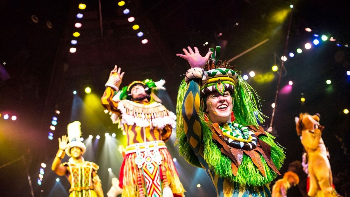 Performers in vibrant, colorful costumes are energetically dancing on a stage, with bright stage lights creating a festive atmosphere.