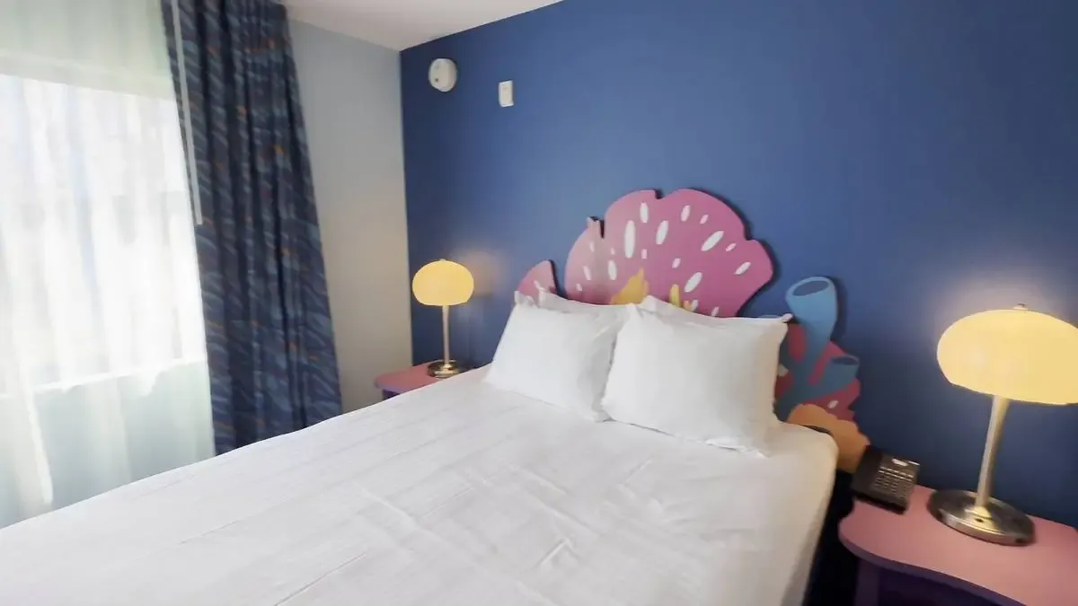 An hotel room featuring a bed with crisp white linens, flanked by two colorful nightstands with round lamps. The wall is painted blue, and a playful, floral-themed headboard adds a touch of whimsy. Natural light filters through the window with patterned curtains.