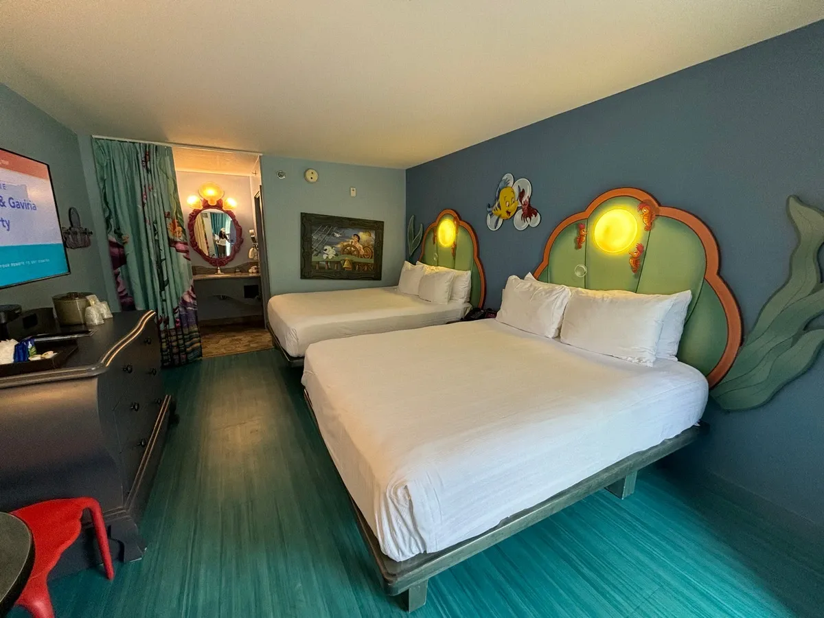 A hotel room with a playful theme features two double beds with shell-shaped headboards and cartoon character decorations on the wall.