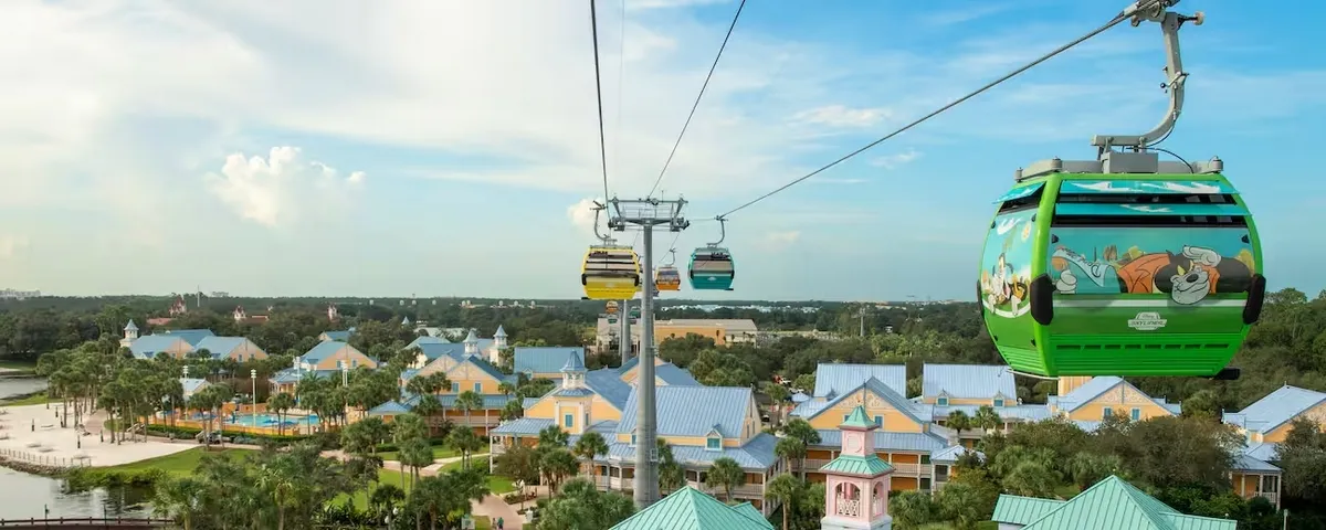 Colorful cable car system over a scenic resort area with buildings painted in pastel colors, palm trees, and a pool, set against a backdrop of greenery and a bright blue sky.
