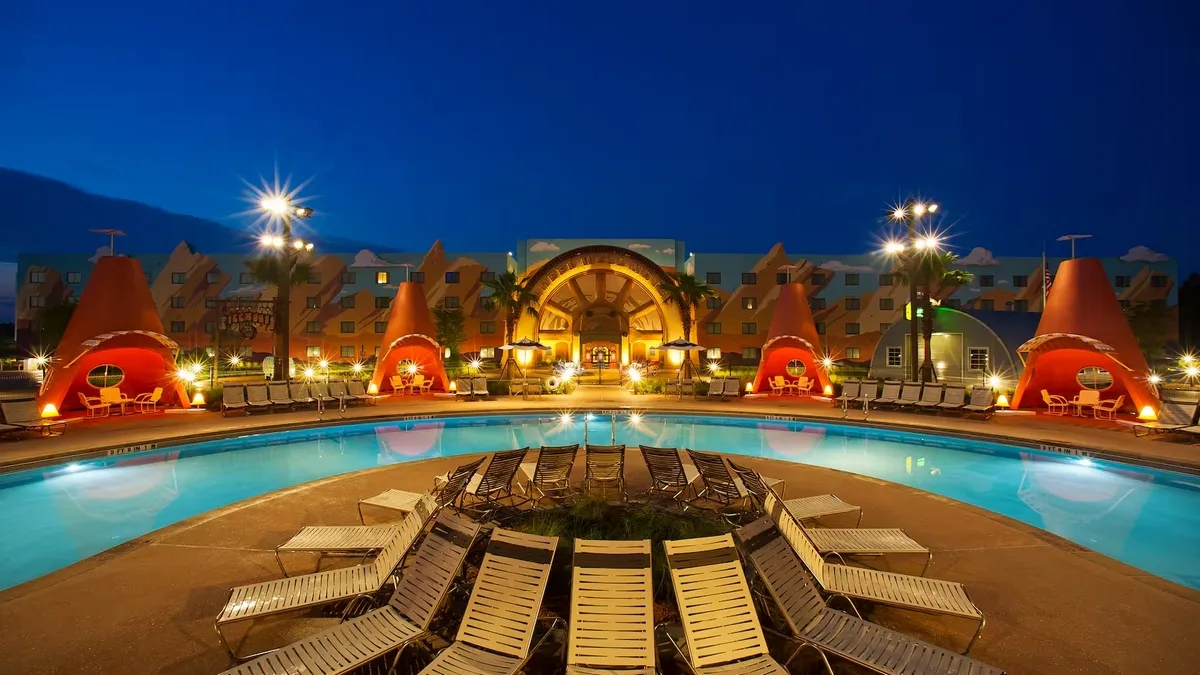 A nighttime scene at a pool area features several cabanas glowing warmly against a deep blue sky. Lounge chairs are arranged around the curved pool, reflecting the lights from the cabanas and nearby palm trees.