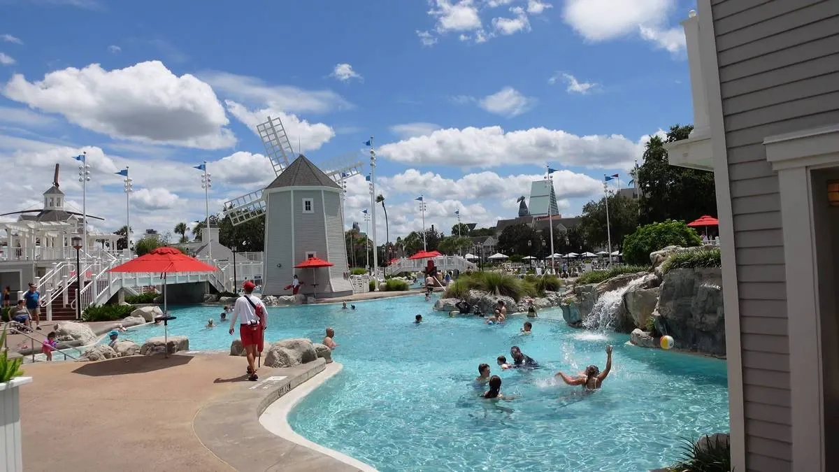 People enjoy a swimming pool with a lifeguard nearby, surrounded by rocks and a small waterfall. There's a windmill structure and red umbrellas around the area.