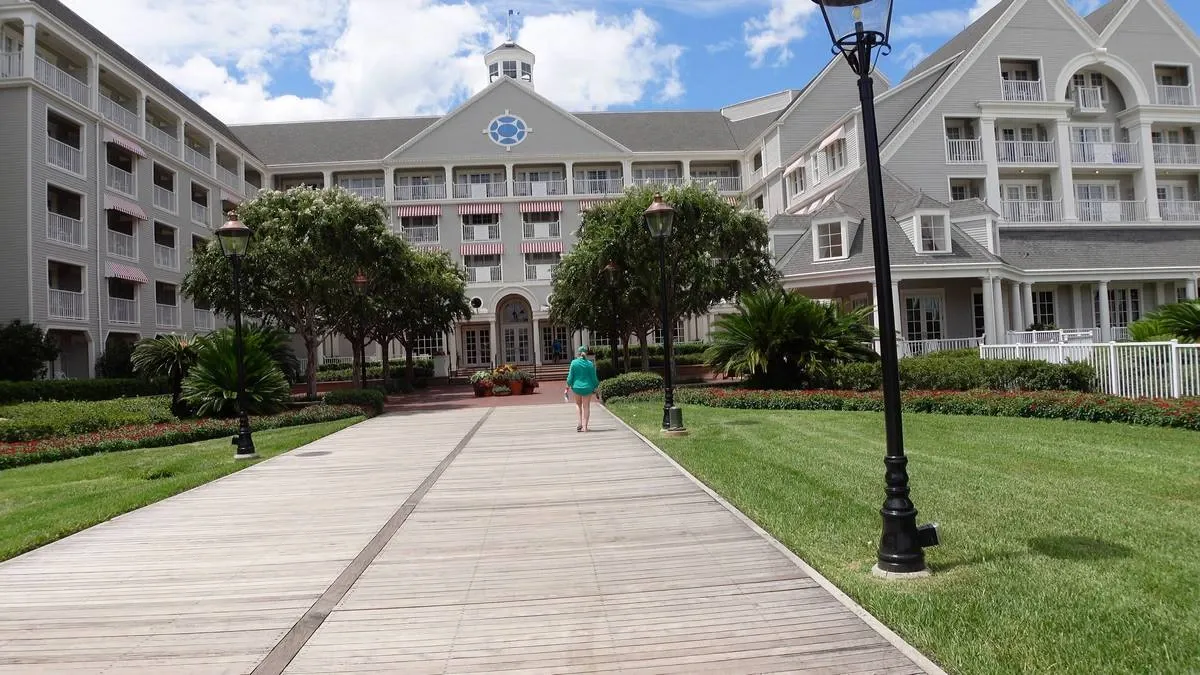 A person walks along a wooden pathway leading to a large, elegant hotel with a gray facade and white trim. The building is surrounded by lawns and trees, with vintage-style lamp posts lining the path.