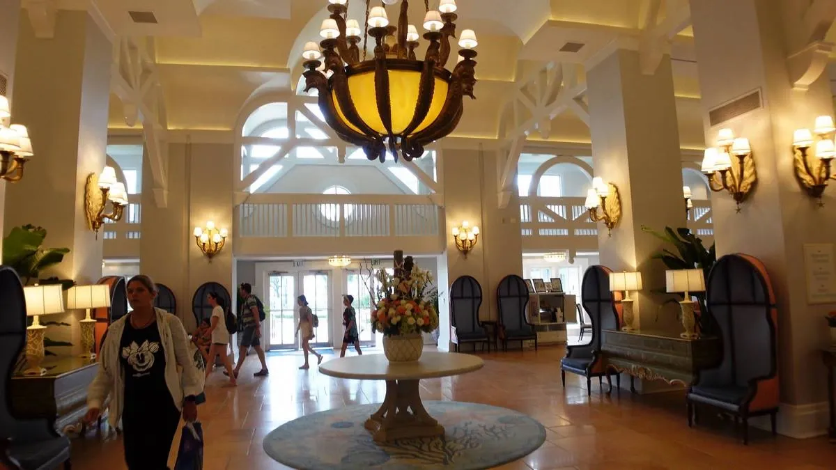 A spacious hotel lobby with a grand chandelier hanging from a high ceiling, featuring elegant wall sconces and plush seating. A round table with a floral arrangement sits in the center, while several people walk through the area.