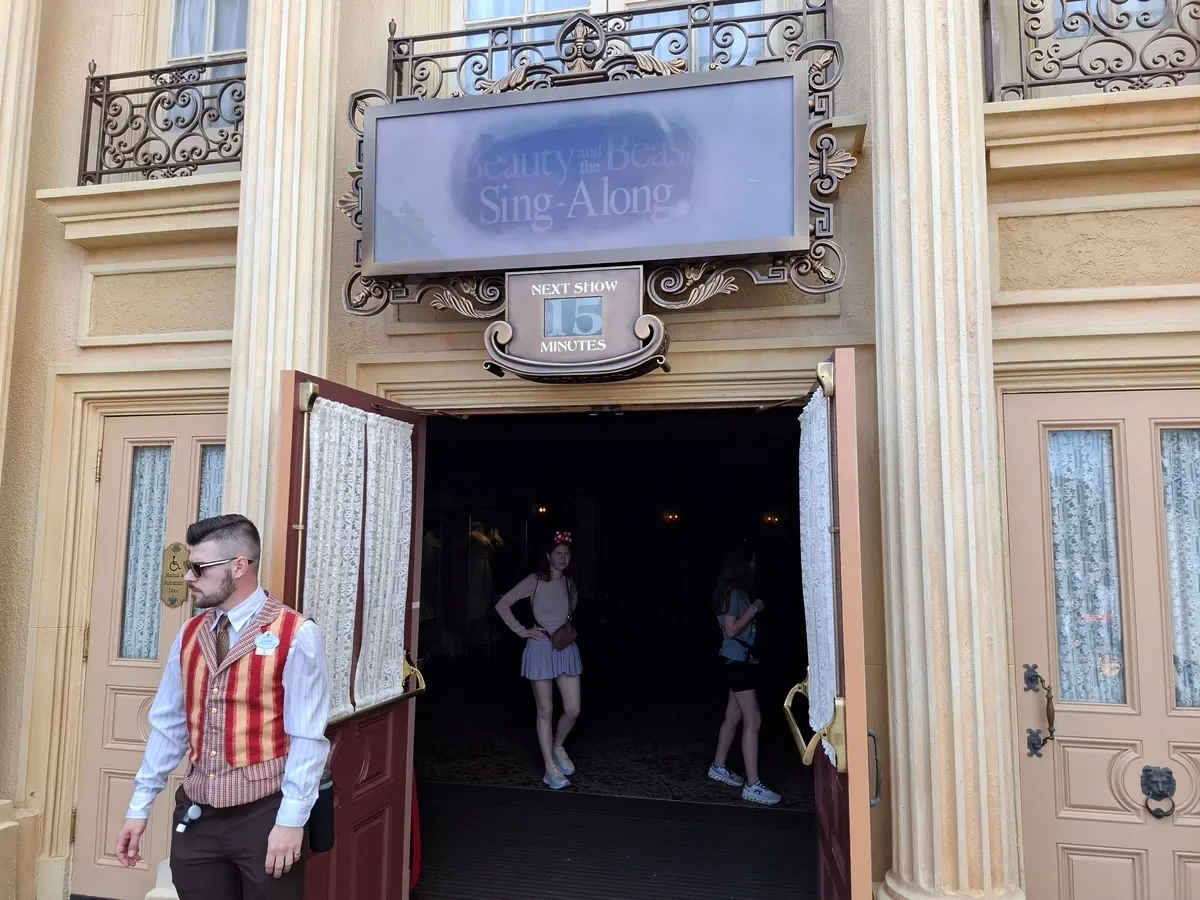 The entrance of a "Beauty and the Beast Sing-Along" attraction, with a sign indicating the next show is in 15 minutes. The entrance features ornate columns and partially open doors, revealing a dimly lit interior where a person with Minnie Mouse ears is visible.