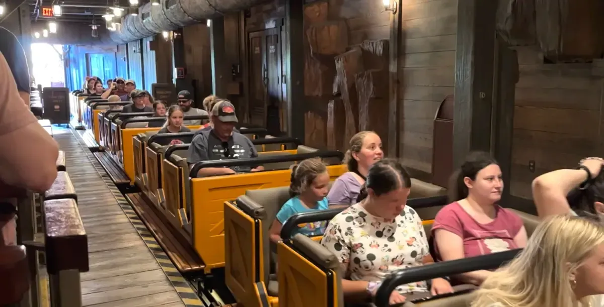 A group of people are seated in a yellow roller coaster train inside a station with rustic decorations, ready for the ride to begin.