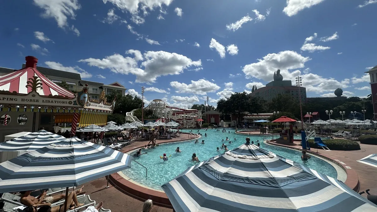 People enjoy a swimming pool surrounded by striped umbrellas. A blue sky with scattered clouds and nearby buildings complete the lively scene.