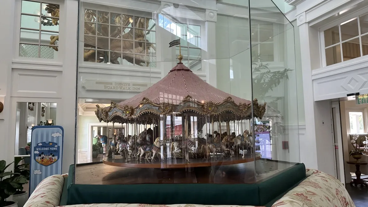 A miniature carousel is encased in glass, displayed in the elegant lobby of Disney's BoardWalk Inn. The bright, welcoming space features white walls and large windows, with a "Welcome Home" sign nearby.