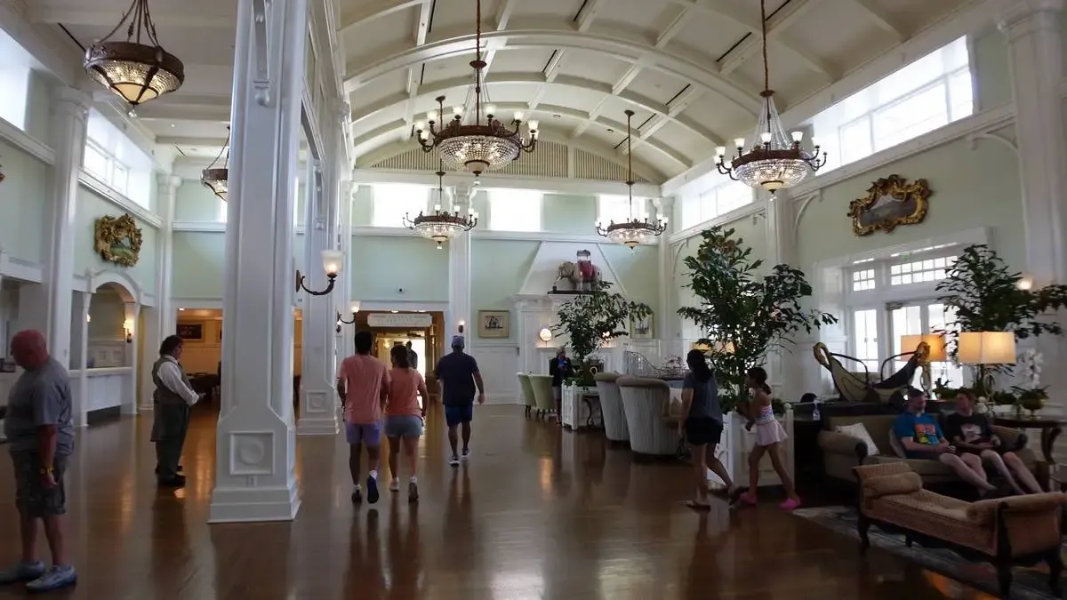 A spacious hotel lobby with high ceilings, elegant chandeliers, and large windows letting in natural light. Several people walk through the area, while others relax on comfortable sofas.