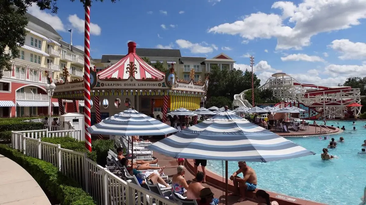 People enjoy a swimming pool surrounded by striped umbrellas. A blue sky with scattered clouds and nearby buildings complete the lively scene.